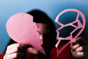 Close-up of hands holding a pink broken paper heart, symbolizing love and heartbreak.