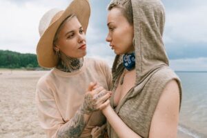 Two women with tattoos, sharing a heartfelt moment on a beach, portraying love and friendship.